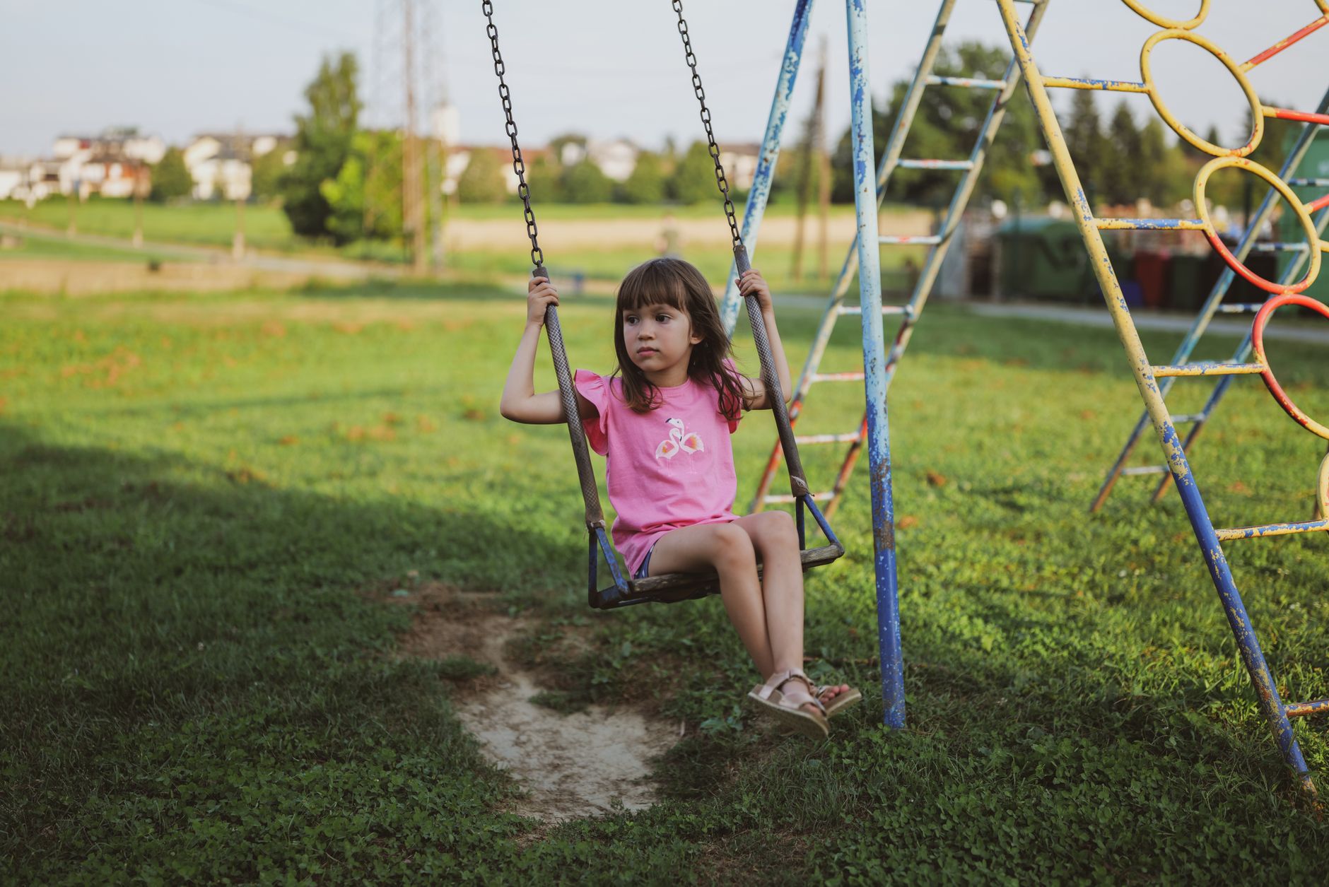 A young girl in a pink shirt swings in a rural playground on a sunny day.